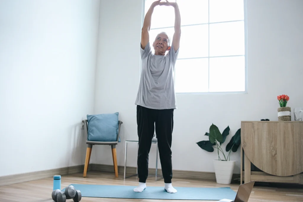 An older man stretching his arms overhead on a yoga mat at home
